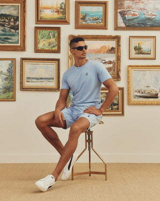 Wearing a Psycho Bunny MENS CLASSIC CREW NECK TEE - B6U014B200 in light blue and sunglasses, a person sits stylishly on a tall stool before a wall of framed landscapes and seascapes, legs crossed with one arm resting on their knee.