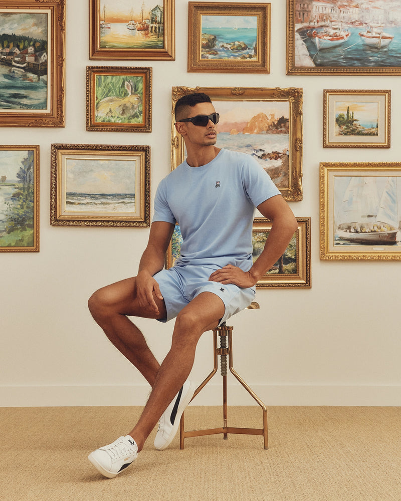Wearing a Psycho Bunny MENS CLASSIC CREW NECK TEE - B6U014B200 in light blue and sunglasses, a person sits stylishly on a tall stool before a wall of framed landscapes and seascapes, legs crossed with one arm resting on their knee.