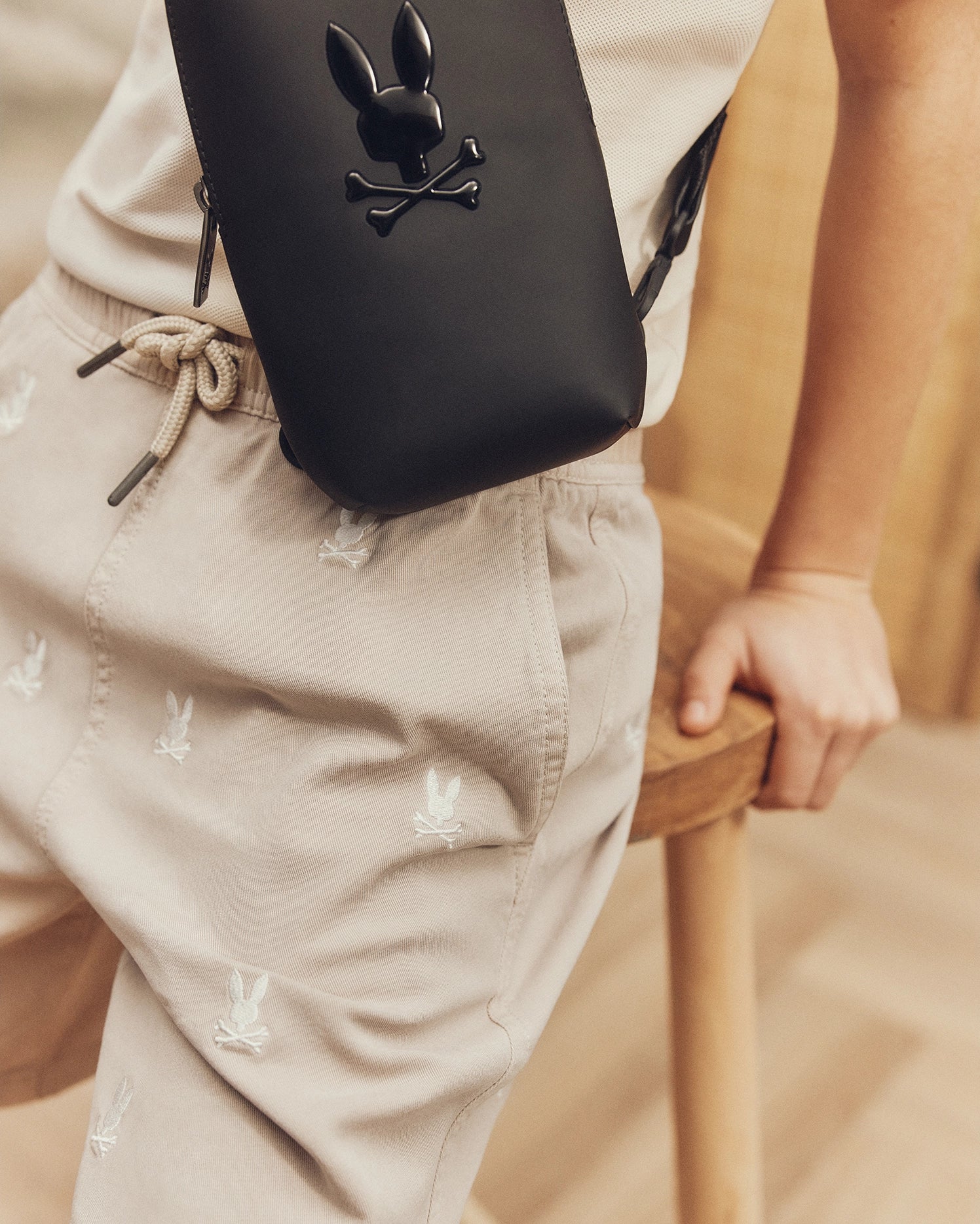 Sporting Psycho Bunny's KIDS ALL OVER WILLIS STRETCH TENCEL SHORT, adorned with white bunny patterns, paired with a crisp white shirt and a crossbody bag flaunting a bunny and crossbones logo. The indoor casual setting enhances their regular fit style effortlessly.