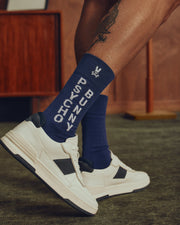 A person with a leg tattoo wears Psycho Bunny MENS 3 PACK DRESS SOCK - B6F504F200 in blue, paired with white sneakers with black accents, standing on a green carpet in a wood-paneled room—a truly stylish ensemble.