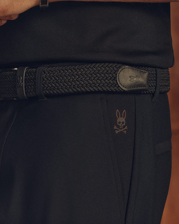 Close-up of a person in stylish black pants with a subtle bunny skull logo, wearing Psycho Bunny's MENS STRETCH BRAIDED BELT (B6A890F200), featuring a small emblem. Their black shirt is neatly tucked into the waistband.