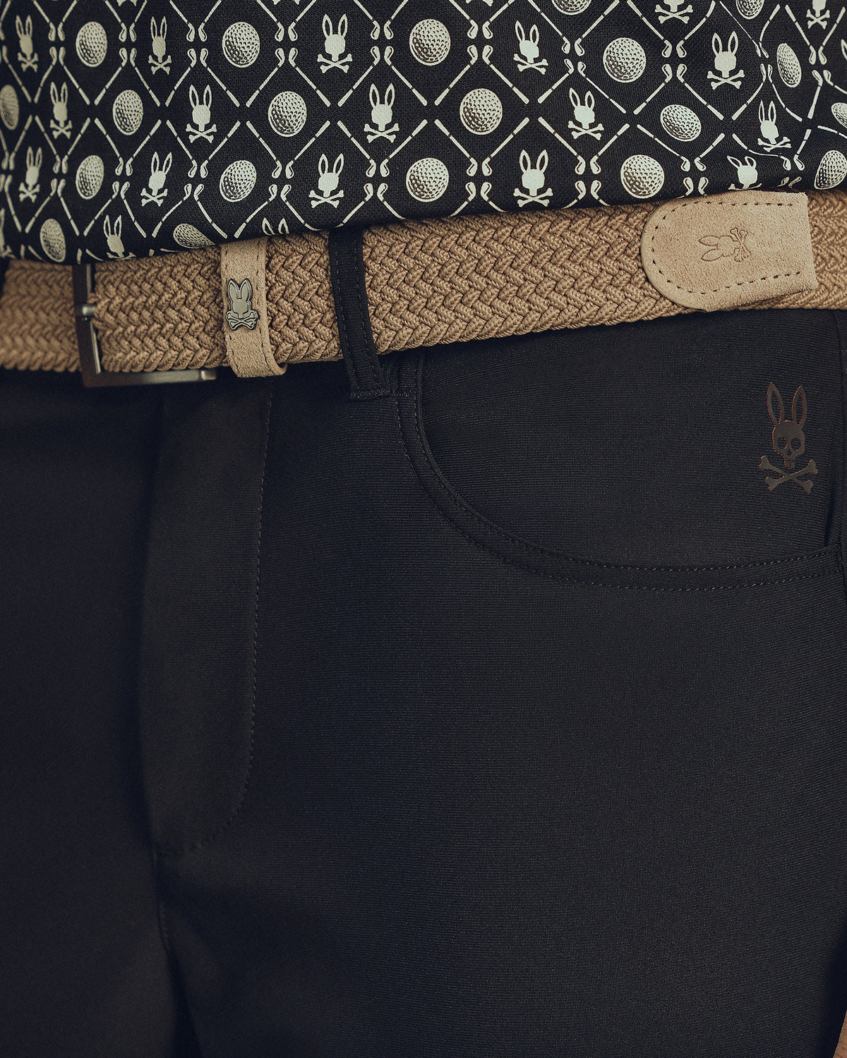Close-up of a waist in dark pants with a skull emblem, featuring Psycho Bunny's MENS STRETCH BRAIDED BELT - B6A890F200 in beige with a rabbit logo. The ensemble is completed by a patterned shirt with tennis balls and rabbits for an eye-catching look.