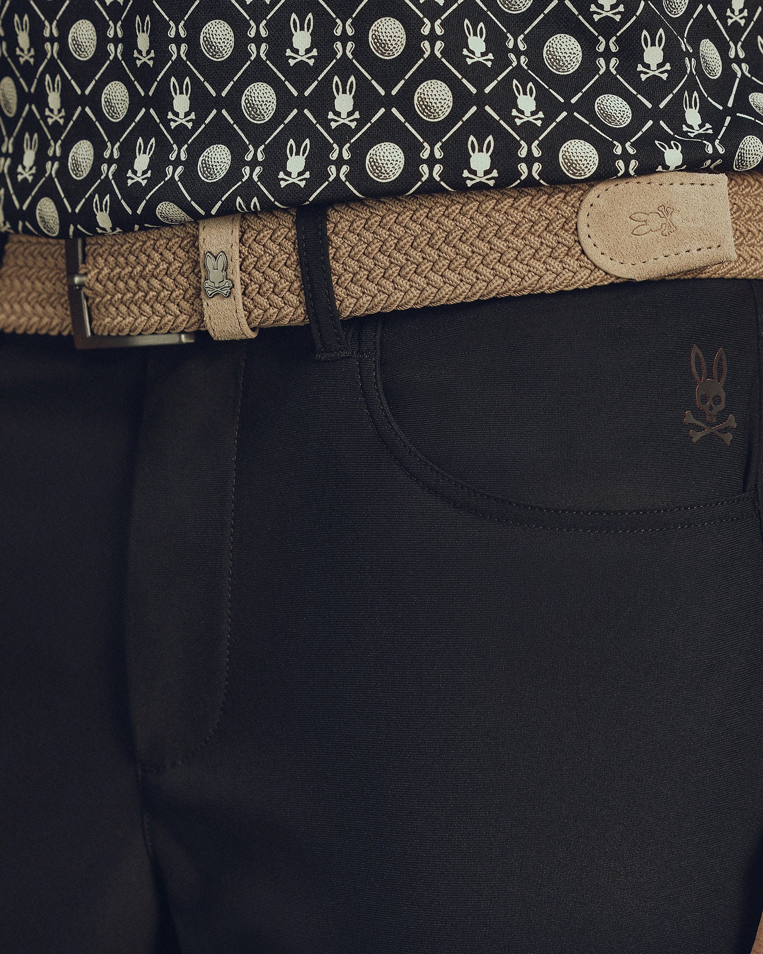 Close-up of a waist in dark pants with a skull emblem, featuring Psycho Bunny's MENS STRETCH BRAIDED BELT - B6A890F200 in beige with a rabbit logo. The ensemble is completed by a patterned shirt with tennis balls and rabbits for an eye-catching look.