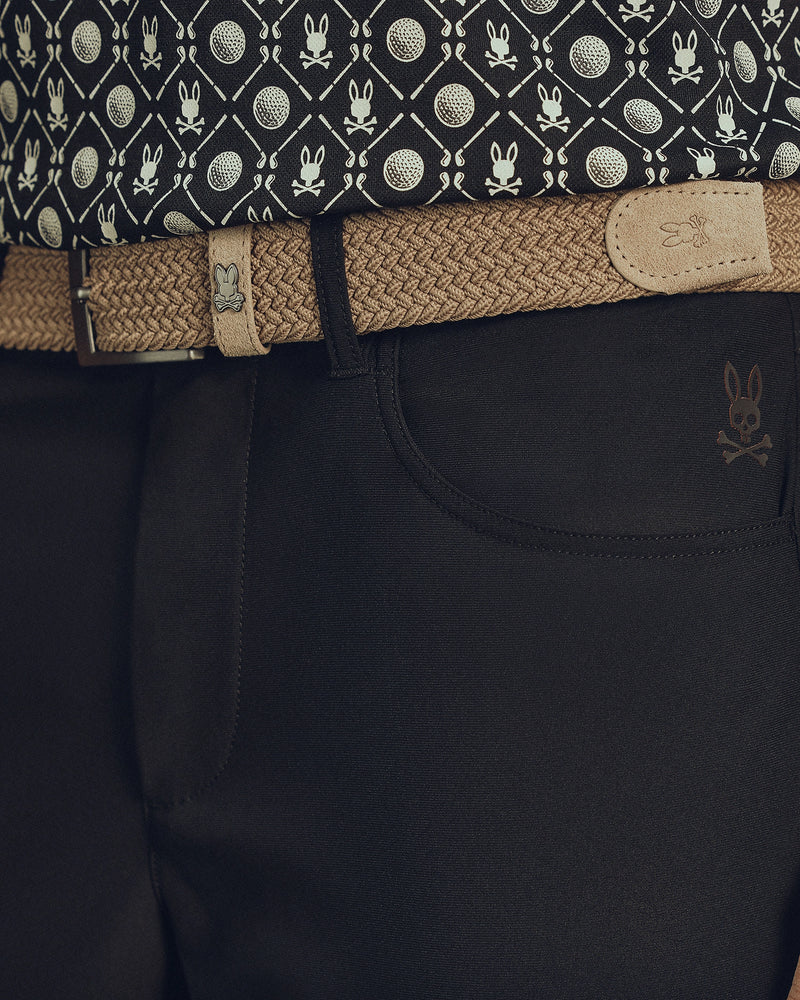 Close-up of a waist in dark pants with a skull emblem, featuring Psycho Bunny's MENS STRETCH BRAIDED BELT - B6A890F200 in beige with a rabbit logo. The ensemble is completed by a patterned shirt with tennis balls and rabbits for an eye-catching look.