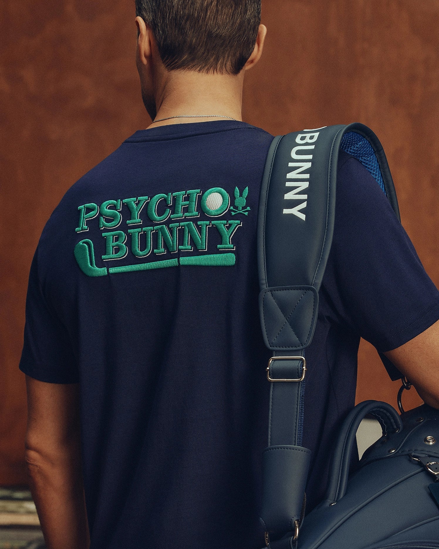 The image shows a short-haired person turned away, wearing the Psycho Bunny MENS DENIS GRAPHIC TEE in navy with green 