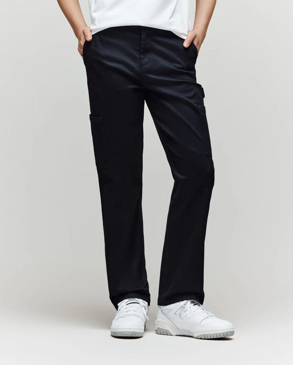A child stands against a plain background, wearing Psycho Bunny KIDS RUSSELL CARPENTER PANT - KB0600001 in black with cargo pockets, paired with a white shirt and white sneakers. Their hands are in their pockets and their face is not shown.