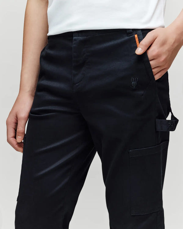 A person wears Psycho Bunny KIDS RUSSELL CARPENTER PANT - KB0600001 in black, with cargo pockets and a small rabbit embroidery near the pocket, styled with a white shirt. The image highlights their waist and upper legs as one hand rests in the pocket.