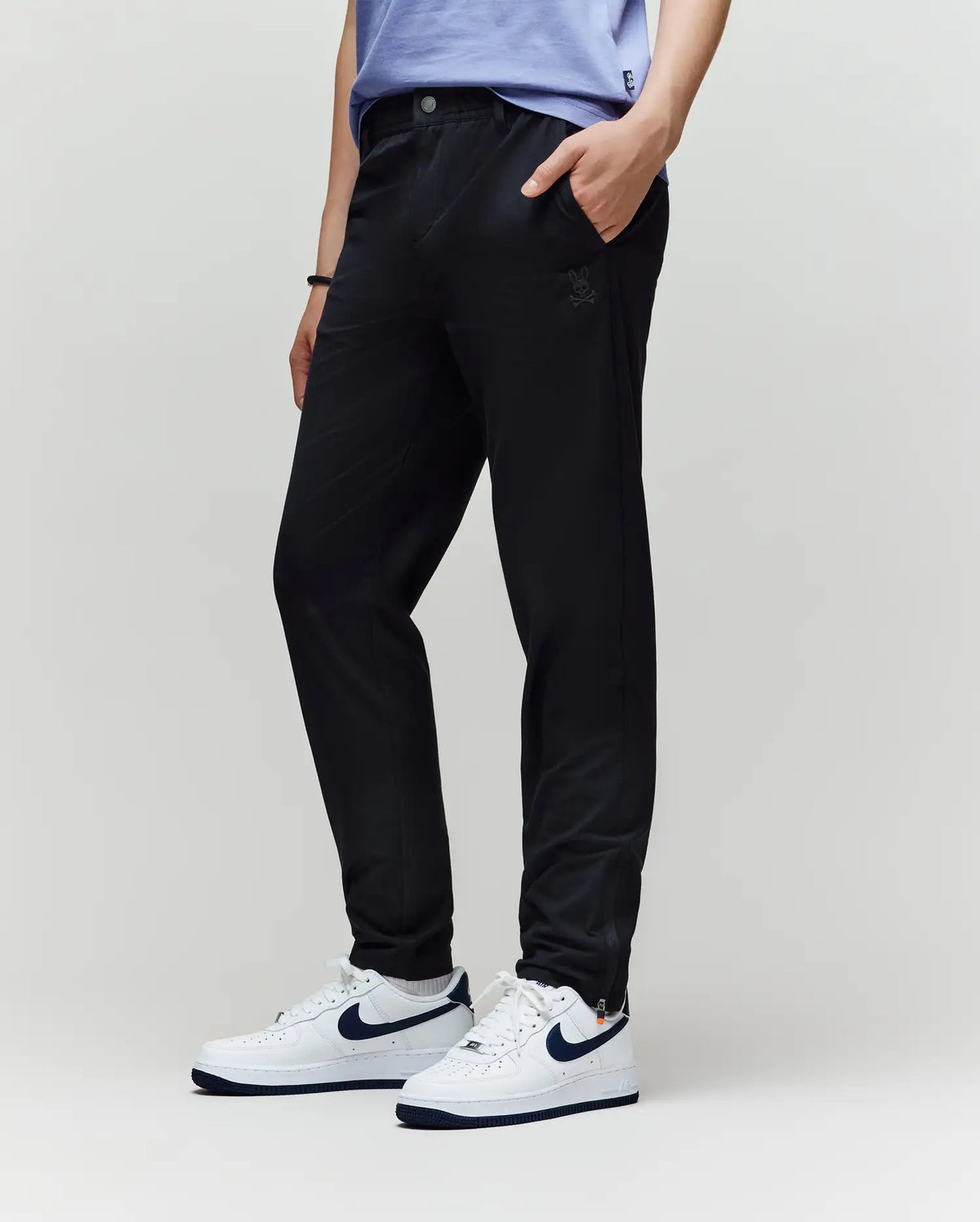 A person in Psycho Bunny KIDS MATHIEU COMMUTER PANT (KB0600002), a light blue shirt, and white Nike sneakers with a dark blue swoosh stands with one hand in their pocket against a plain light background.
