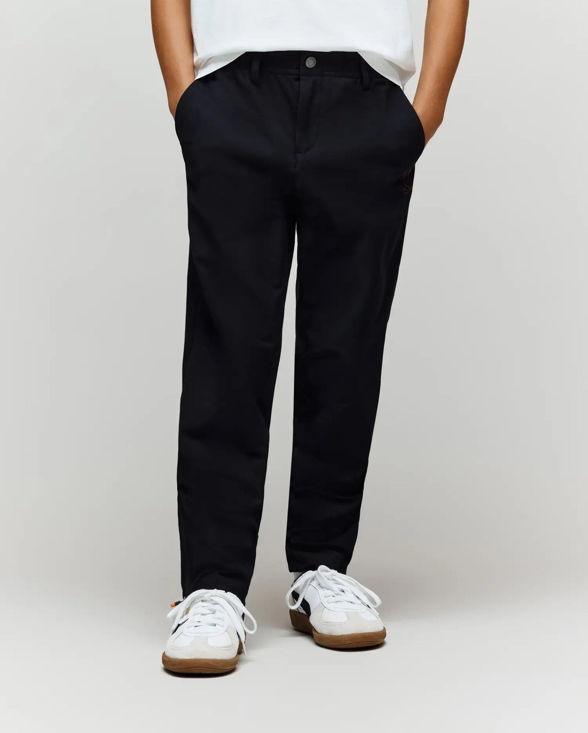 A child wears Psycho Bunny’s KIDS MATHIEU COMMUTER PANT (KB0600002) in classic black with subtle Bunny embroidery, paired with white sneakers, hands in pockets. Only the lower half is shown against a plain light background.