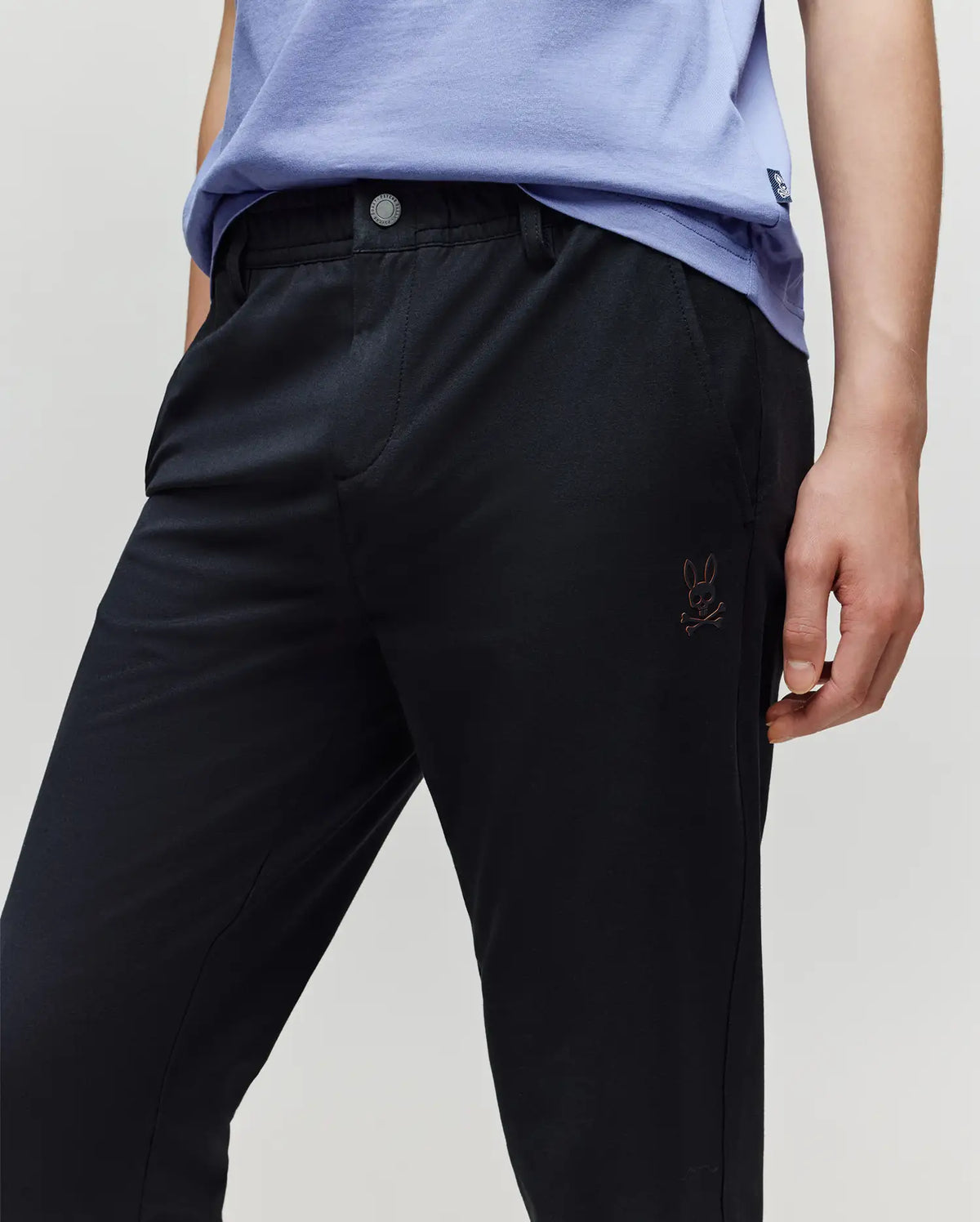 A child wears a light purple shirt and Psycho Bunny KIDS MATHIEU COMMUTER PANT (KB0600002), styled with one hand in the pocket; signature Bunny embroidery appears near the left pocket.
