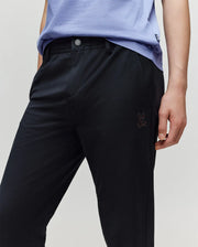 A child wears a light purple shirt and Psycho Bunny KIDS MATHIEU COMMUTER PANT (KB0600002), styled with one hand in the pocket; signature Bunny embroidery appears near the left pocket.