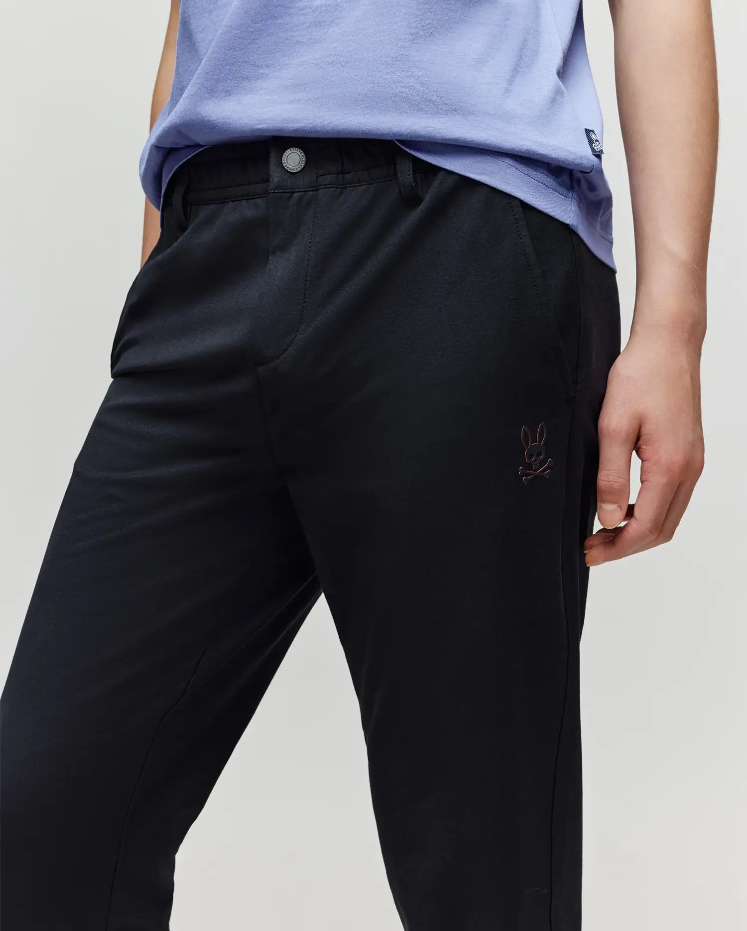 A child wears a light purple shirt and Psycho Bunny KIDS MATHIEU COMMUTER PANT (KB0600002), styled with one hand in the pocket; signature Bunny embroidery appears near the left pocket.