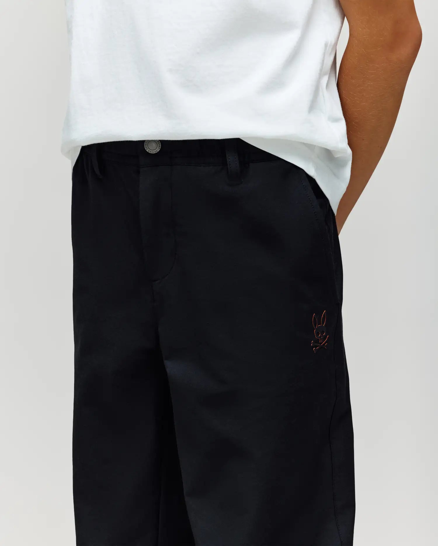A child wears Psycho Bunny's KIDS MATHIEU COMMUTER PANT (KB0600002) with signature Bunny embroidery on the pocket and a white shirt, standing with hands in pockets.