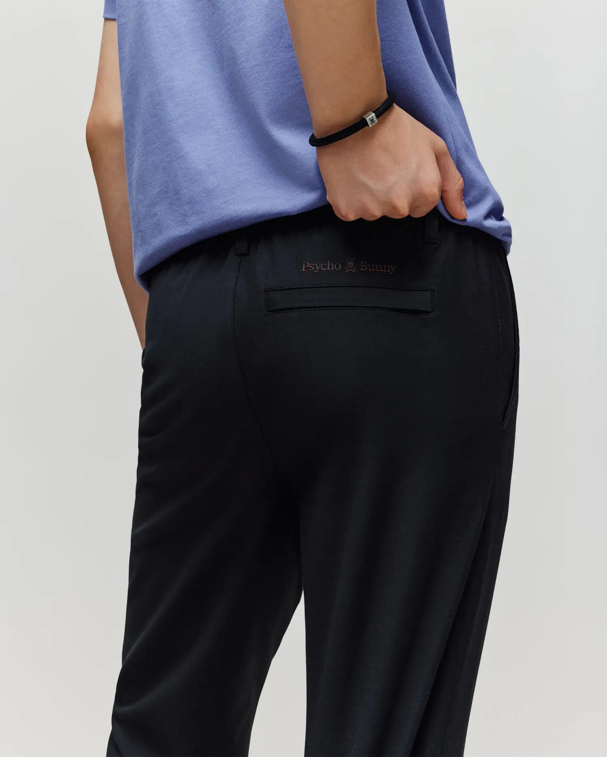 A person in a light purple shirt and black Psycho Bunny KIDS MATHIEU COMMUTER PANT (KB0600002) is shown from behind, one hand in their pocket; the classic fit pants feature signature Bunny embroidery above the back pocket.