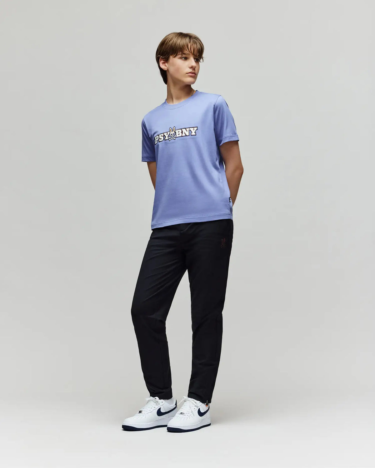 A young person stands against a plain background in Psycho Bunny's KIDS MATHIEU COMMUTER PANT (KB0600002), a lavender "PSYBNY" T-shirt, and white Nike sneakers with black swooshes, glancing sideways with hands behind their back.