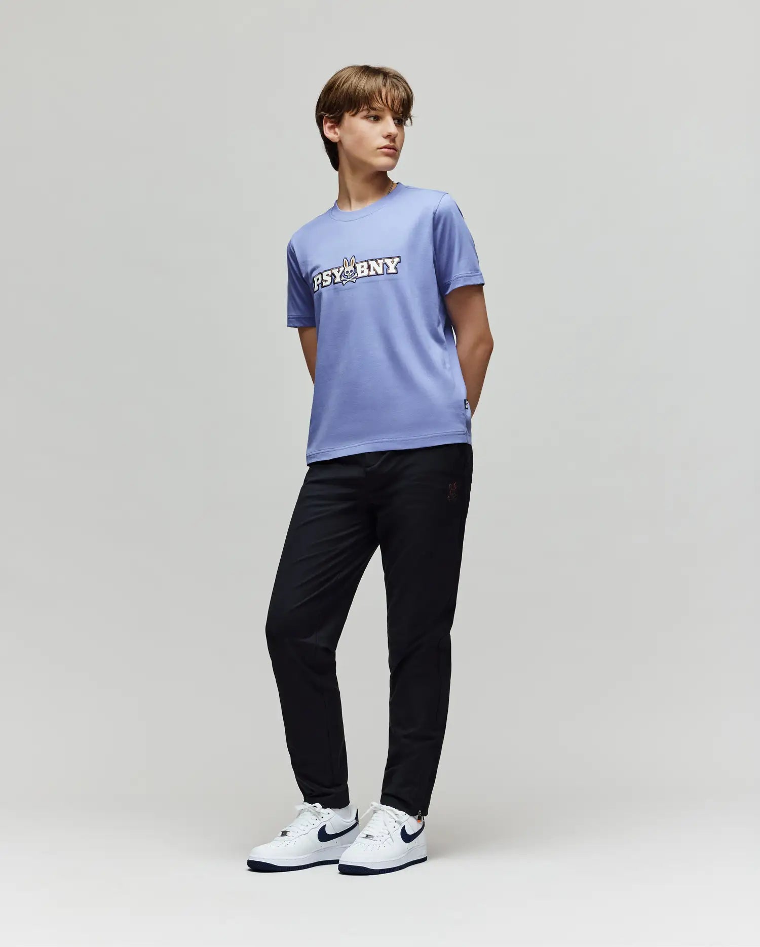 A young person stands against a plain background in Psycho Bunny's KIDS MATHIEU COMMUTER PANT (KB0600002), a lavender "PSYBNY" T-shirt, and white Nike sneakers with black swooshes, glancing sideways with hands behind their back.