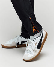 A close-up of a person in Psycho Bunny KIDS MATHIEU COMMUTER PANT (KB0600002) featuring an orange skull graphic inside the ankle zipper, paired with white and black Puma sneakers with gum soles.