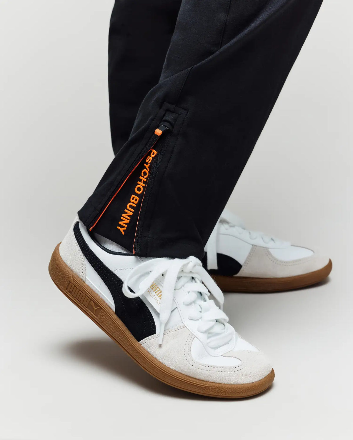 A person wearing Psycho Bunny KIDS MATHIEU COMMUTER PANT (KB0600002), classic fit black pants with Bunny embroidery and an orange tag, paired with white Puma sneakers with black stripes and gum soles, shown from the ankle down.