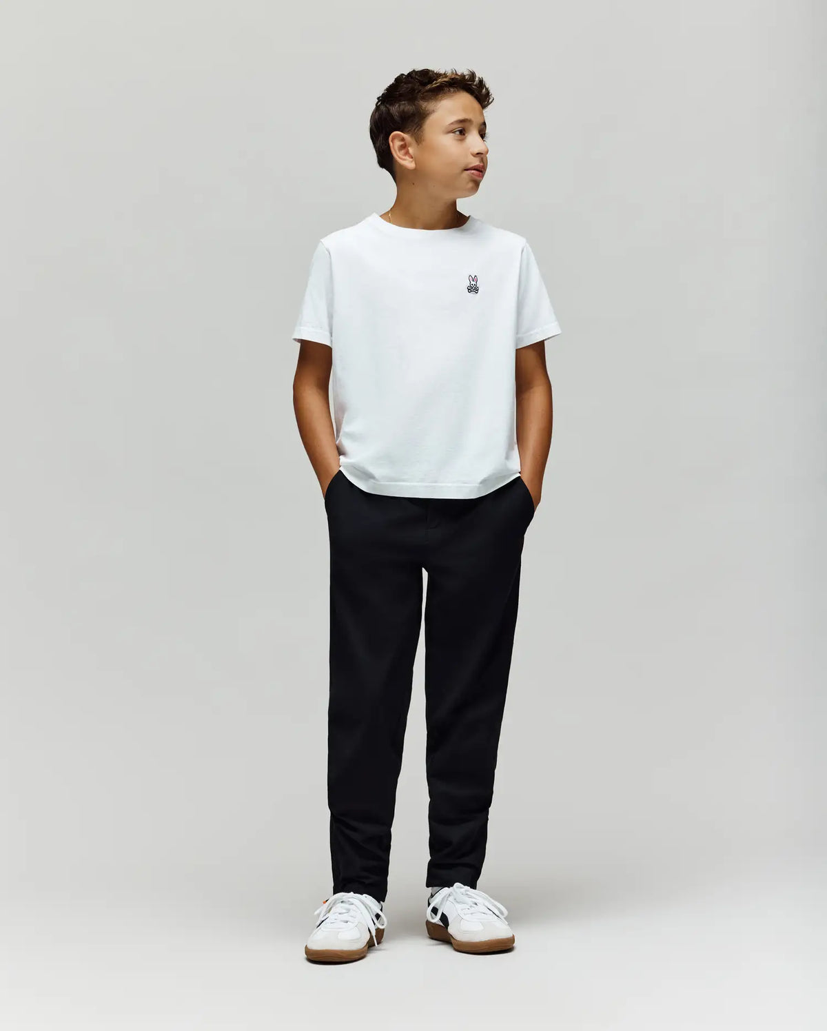 A boy stands against a plain light gray background, wearing a white t-shirt, Psycho Bunny KIDS MATHIEU COMMUTER PANT (KB0600002), and white sneakers. His hands are in his pockets, looking to the side.