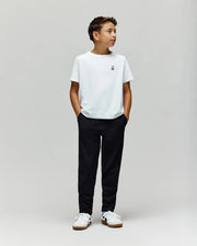 A boy stands against a plain light gray background, wearing a white t-shirt, Psycho Bunny KIDS MATHIEU COMMUTER PANT (KB0600002), and white sneakers. His hands are in his pockets, looking to the side.