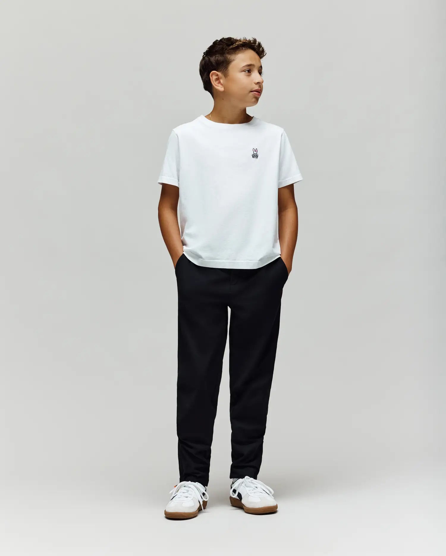 A boy stands against a plain light gray background, wearing a white t-shirt, Psycho Bunny KIDS MATHIEU COMMUTER PANT (KB0600002), and white sneakers. His hands are in his pockets, looking to the side.