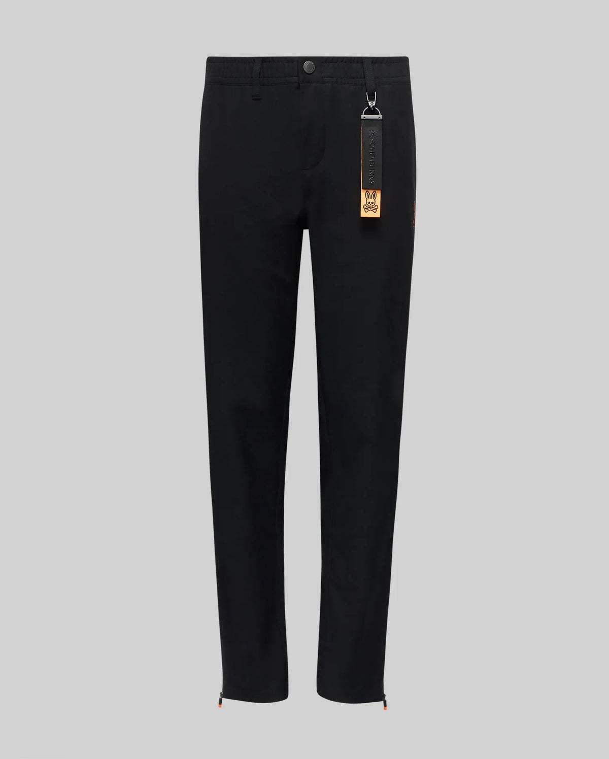 The Psycho Bunny KIDS MATHIEU COMMUTER PANT (KB0600002) features a black straight-leg, classic fit with waistband, button and front zipper. A black tag with keychain and orange label hangs from a belt loop. Shown on a light gray background.