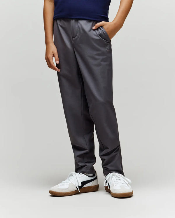 A person wears Psycho Bunny KIDS MATHIEU COMMUTER PANT KB0600002 in classic gray with subtle bunny embroidery, paired with white sneakers and a dark shirt. They stand with one hand in their pocket against a plain, light background.