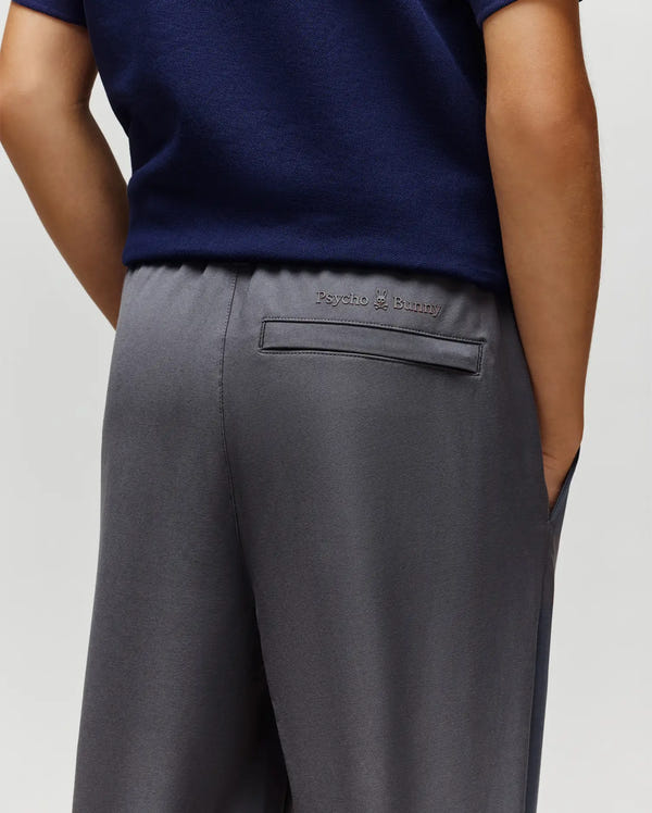 A person wears a navy blue shirt and Psycho Bunny KIDS MATHIEU COMMUTER PANT (KB0600002) in classic gray fit, one hand in a pocket. Signature Bunny embroidery is visible above the back pocket.