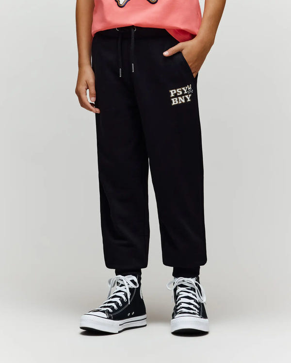 A child wears Psycho Bunny’s KIDS WADE CORD PATCH SWEATPANT (KB0800002) in black, featuring an embroidered "PSY BN" wordmark on the left thigh. Only the lower half is visible, paired with a coral tee and black high-top sneakers against a plain background.