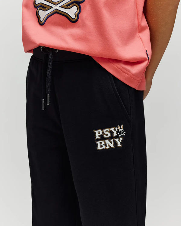 A person wears a pink tee with a skull and crossbones graphic and Psycho Bunny’s KIDS WADE CORD PATCH SWEATPANT (KB0800002), black sweatpants featuring an embroidered “PSY BNY” wordmark and small rabbit logo, for a classic athletic look.