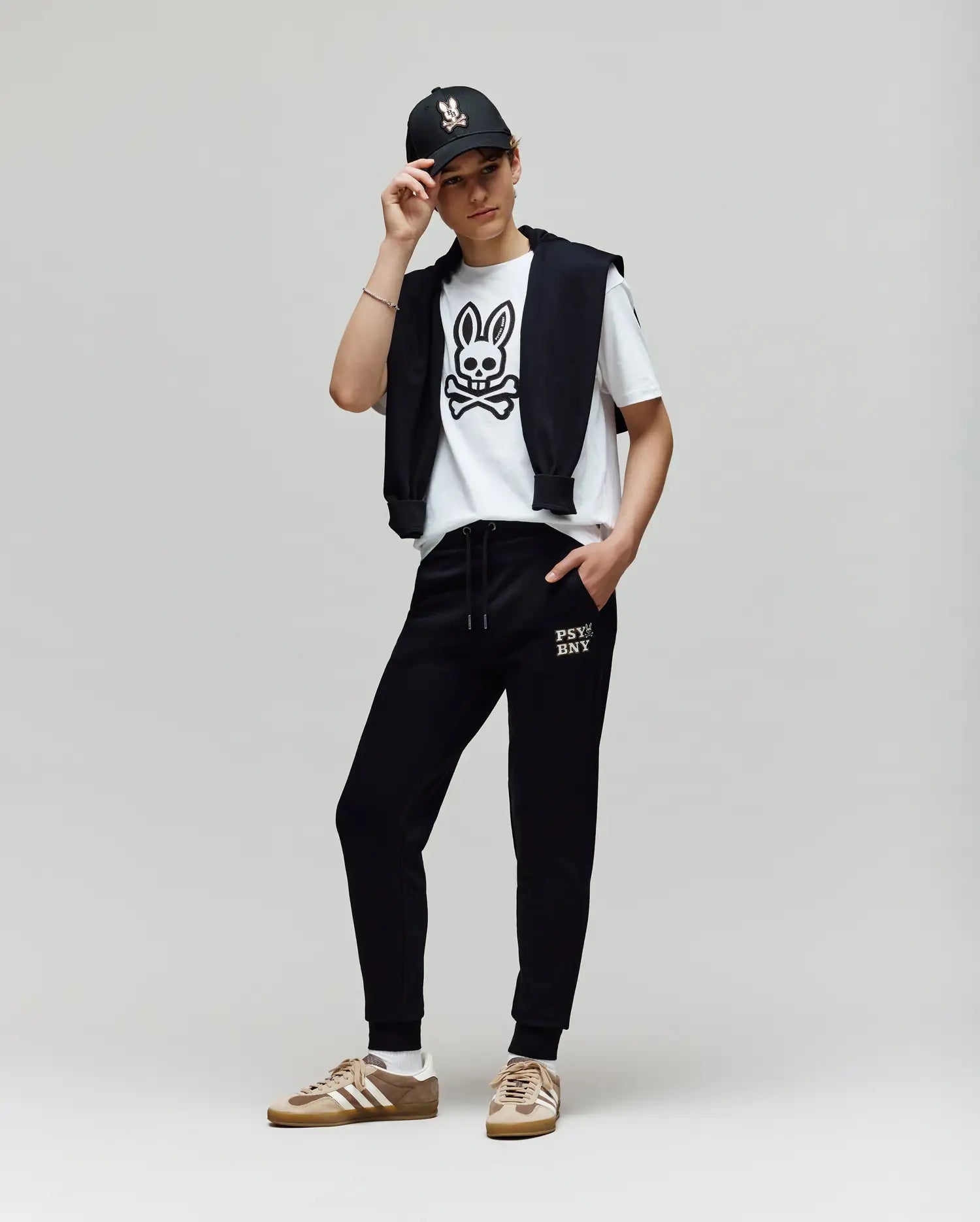 A person poses against a plain background, wearing a black cap, a white Psycho Bunny T-shirt with a bold cartoon bunny graphic, KIDS WADE CORD PATCH SWEATPANT - KB0800002 by Psycho Bunny, and tan Adidas sneakers.