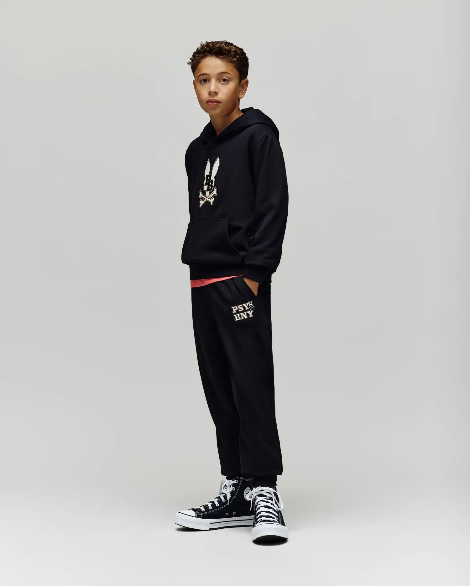 A boy with short curly hair stands against a plain background wearing Psycho Bunny KIDS WADE CORD PATCH SWEATPANT (KB0800002), a black hoodie with a bunny graphic, and black high-top sneakers, hands in his pockets.