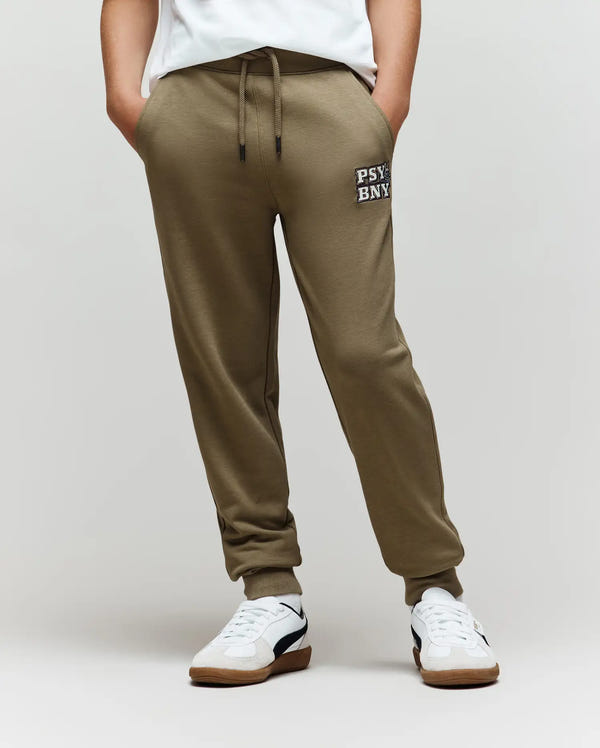 A person wears Psycho Bunny KIDS WADE CORD PATCH SWEATPANT (KB0800002) in olive green with a drawstring waist and "PSY-BNY" on the left thigh, paired with white sneakers, a white shirt, hands in pockets, against a plain background.