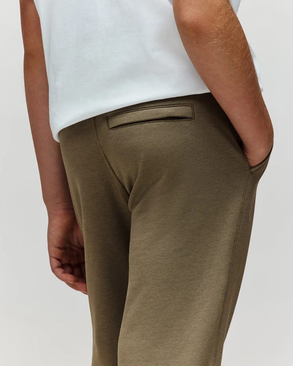 A child wears Psycho Bunny KIDS WADE CORD PATCH SWEATPANT (KB0800002) in olive green with a drawstring waist and a white t-shirt, standing with one hand in their pocket; only the lower back and arm are visible.