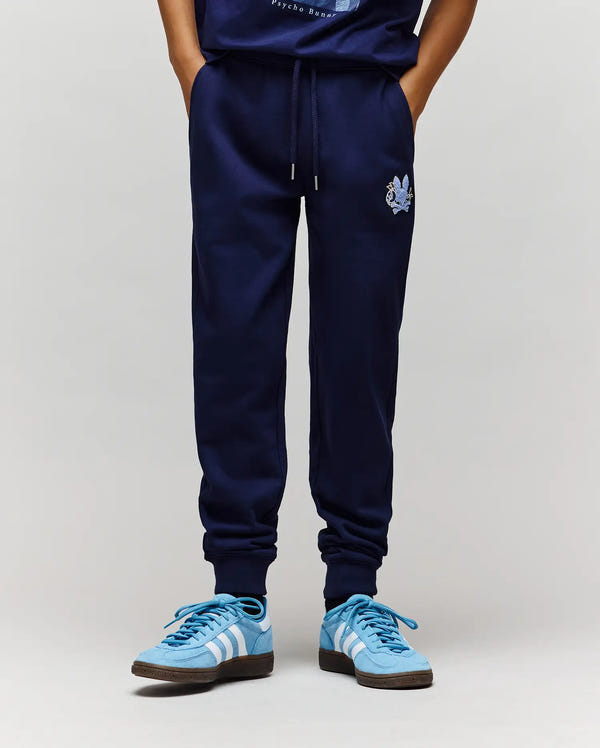 A person stands against a plain background wearing Psycho Bunny KIDS AUGUSTE JOGGER - KB0800003, crafted from 100% cotton with a small embroidered graphic on the left thigh, paired with light blue Adidas sneakers.