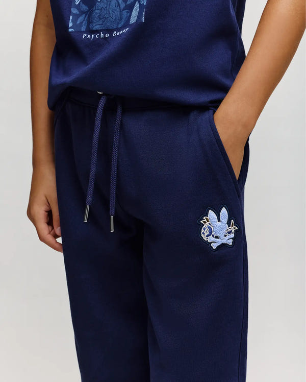 A child wears Psycho Bunny’s KIDS AUGUSTE JOGGER (KB0800003) in navy, 100% cotton, with a drawstring, chenille patches, and an embroidered cartoon rabbit face on the left thigh, right hand in the pocket.
