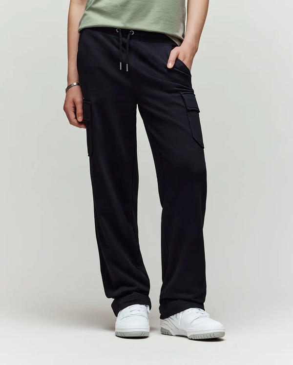 A person models Psycho Bunny’s KIDS NASH CARGO SWEATPANT (KB0800004) in black, styled with a light green t-shirt, white sneakers, and a bracelet against a plain background. Focus highlights the cargo jogger’s pockets and comfort.