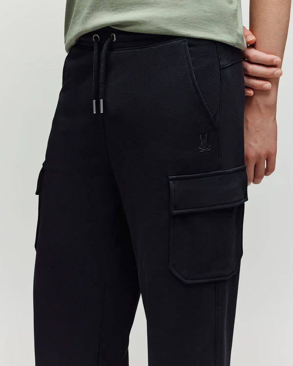A person is shown from the waist down wearing Psycho Bunny KIDS NASH CARGO SWEATPANT - KB0800004 in black, featuring large cargo pockets, an elastic drawstring waistband, and a small embroidered logo, paired with a light green shirt.