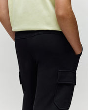 A person wearing a light yellow shirt and Psycho Bunny KIDS NASH CARGO SWEATPANT - KB0800004 stands with one hand in their pocket, shown from the waist down.