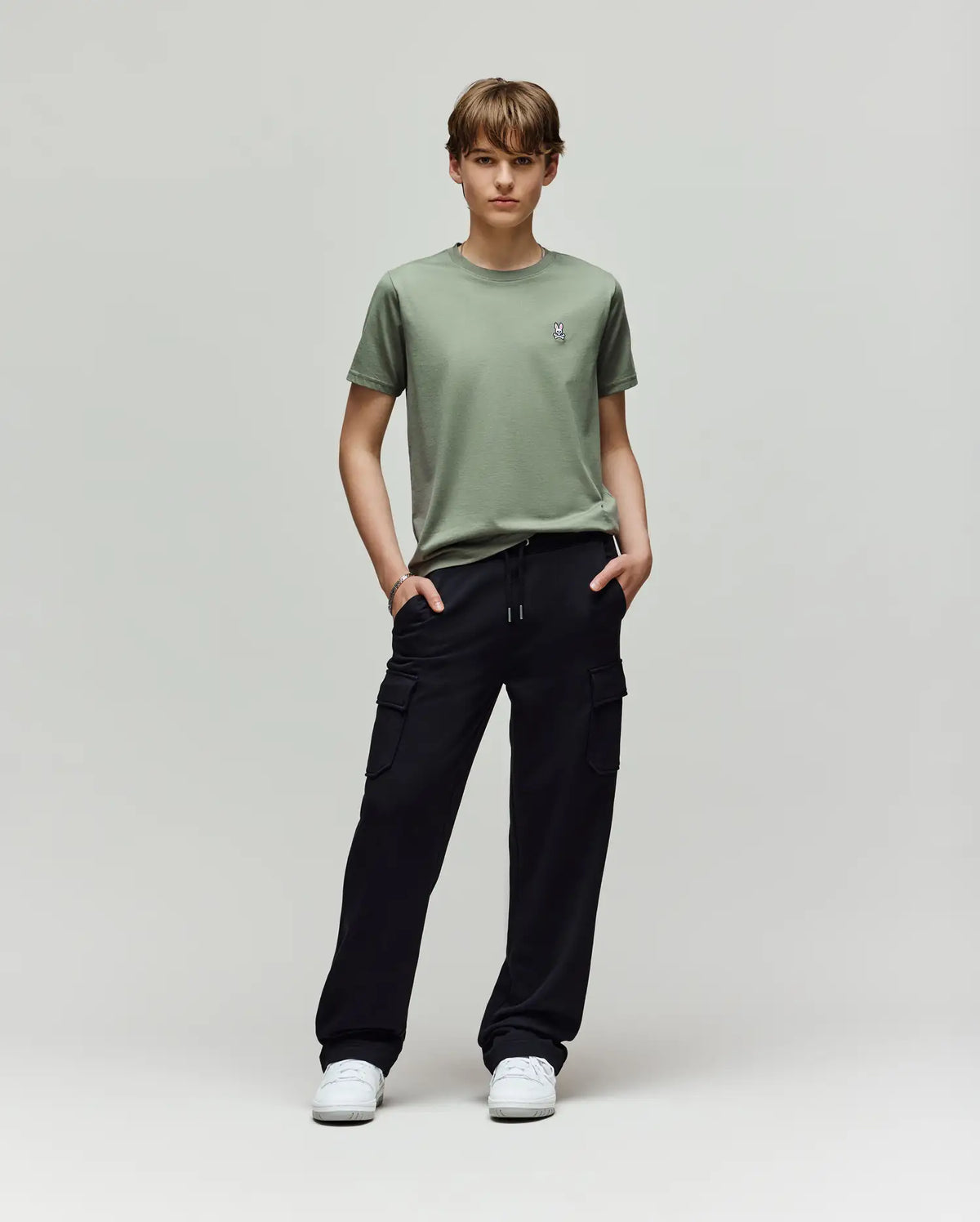A young person faces forward, wearing a light green t-shirt, white sneakers, and Psycho Bunny KIDS NASH CARGO SWEATPANT - KB0800004 with cargo pockets, hands in pockets, enjoying sweatpant comfort against a plain light gray background.