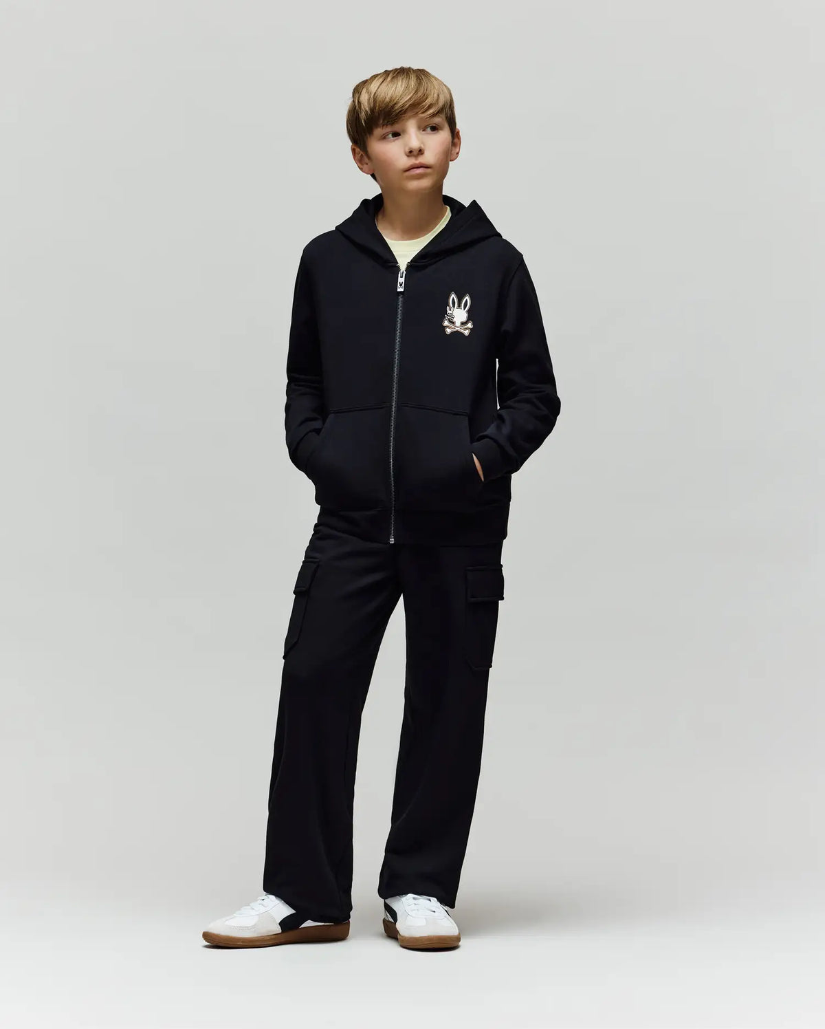 A boy stands with hands in pockets, wearing a black zip-up hoodie with a cartoon chest print, Psycho Bunny KIDS NASH CARGO SWEATPANT - KB0800004 with cargo pockets, and white sneakers, against a plain light grey background.