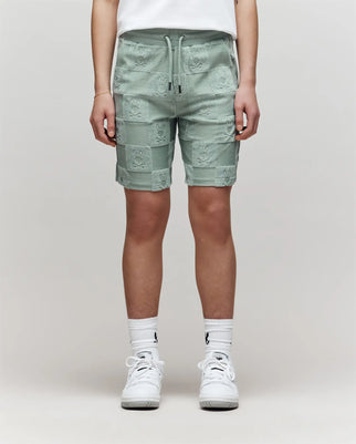 The lower half of a person is shown wearing Psycho Bunny KIDS KELVIN TOWEL TERRY SWEATSHORT - KB0900002 in mint green, paired with white crew socks and sneakers against a plain background.