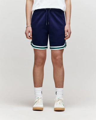 A child models Psycho Bunny’s KIDS BASIL TAPED SHORT (KB0900003) featuring a premium cotton-modal blend and white-green striped hem, paired with white crew socks and sneakers. Only the lower half is shown against a plain light background.
