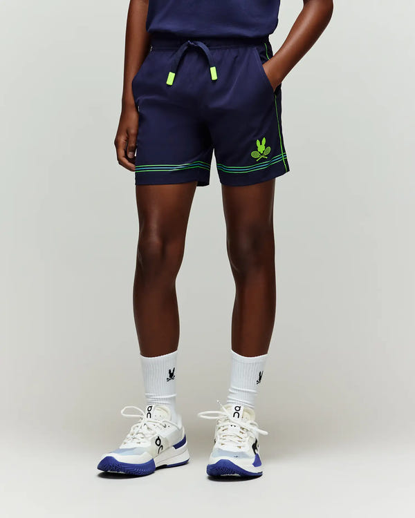 A person is wearing Psycho Bunny KIDS BALL CREW YOUTH SHORT - KB0900011 in navy with green accents, white socks featuring a dark logo, and white and blue athletic shoes, standing against a plain light background.
