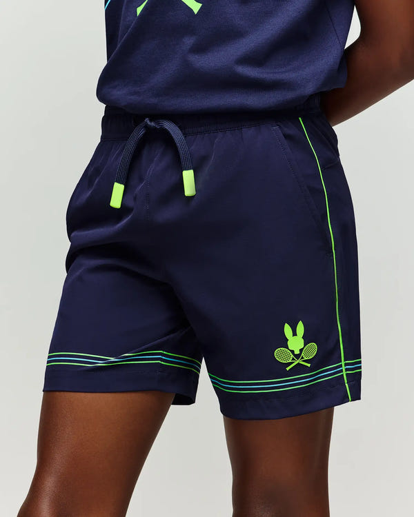 A child wears Psycho Bunny KIDS BALL CREW YOUTH SHORT - KB0900011: navy blue shorts with neon green trim, drawstring waist, and a bunny holding two crossed tennis rackets logo on the left leg.