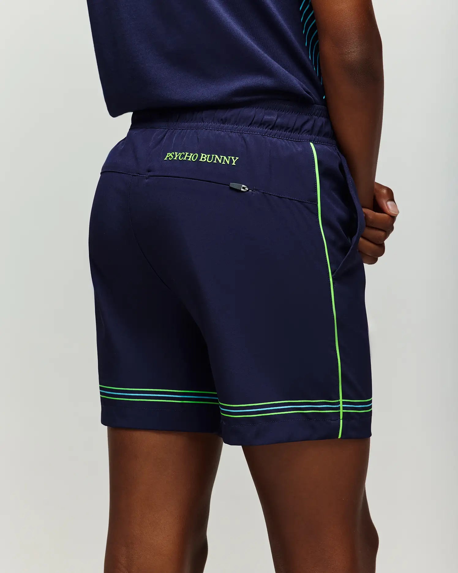 A person wears Psycho Bunny KIDS BALL CREW YOUTH SHORT - KB0900011 in navy with green trim and "Psycho Bunny" on the back waistband, hand in right pocket, standing against a plain background.