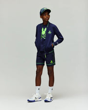 A boy wearing Psycho Bunny KIDS BALL CREW YOUTH SHORT - KB0900011, a navy jacket, tennis graphic tee, matching shorts and cap with neon green accents and Adidas logos, plus white socks and white-blue sneakers.