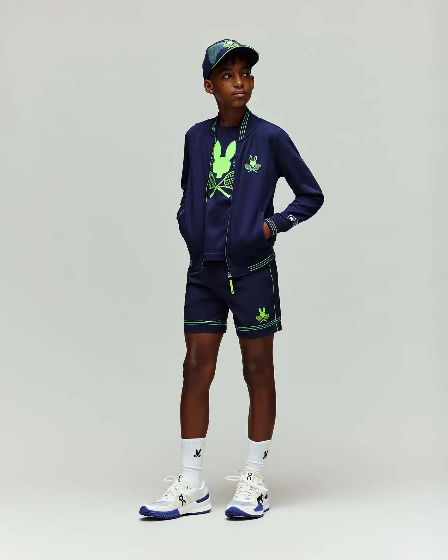 A boy wearing Psycho Bunny KIDS BALL CREW YOUTH SHORT - KB0900011, a navy jacket, tennis graphic tee, matching shorts and cap with neon green accents and Adidas logos, plus white socks and white-blue sneakers.
