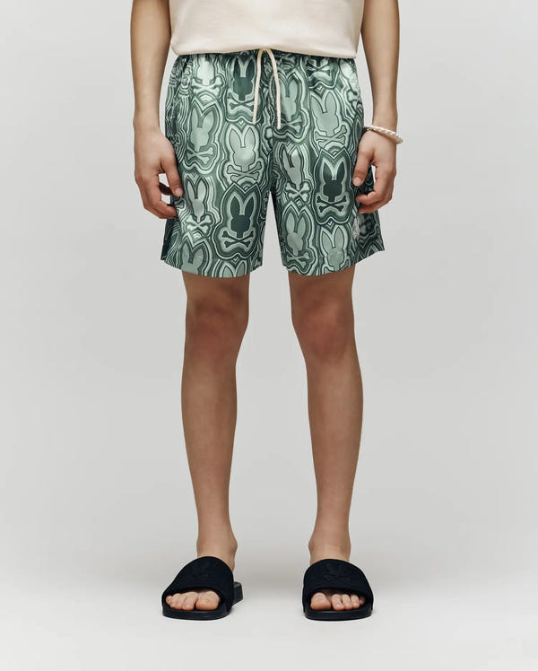 Kids | A person wears Psycho Bunny KIDS CHUCK ALL OVER PRINT LIGHTWEIGHT SWIM TRUNK - KB1000002, featuring abstract bunny faces, paired with a cream shirt, black slide sandals, and a white beaded bracelet against a light background.