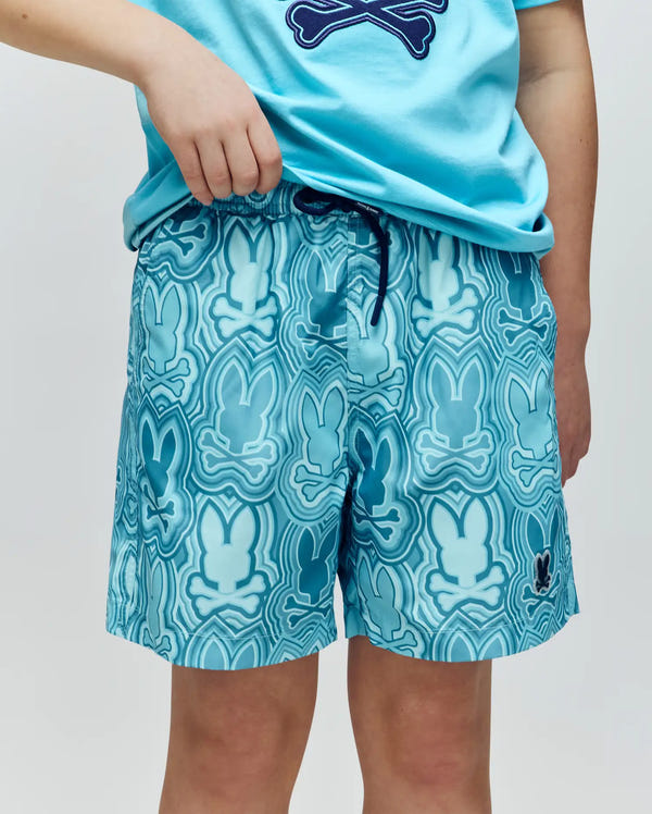 A person wears Psycho Bunny KIDS CHUCK ALL OVER PRINT LIGHTWEIGHT SWIM TRUNK (KB1000002), featuring a wavy dark blue and white bunny print, quick-dry fabric, and a drawstring waistband, paired with a matching blue t-shirt.